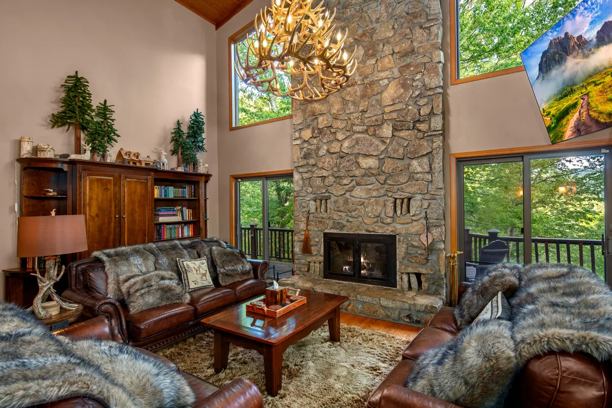 A cozy living space highlighted by a dramatic stone fireplace, antler chandelier, and plush leather sofas arranged for group comfort. Giant windows let in abundant light and showcase beautiful nature views, while decorative plants and warm accents add to the homely atmosphere.