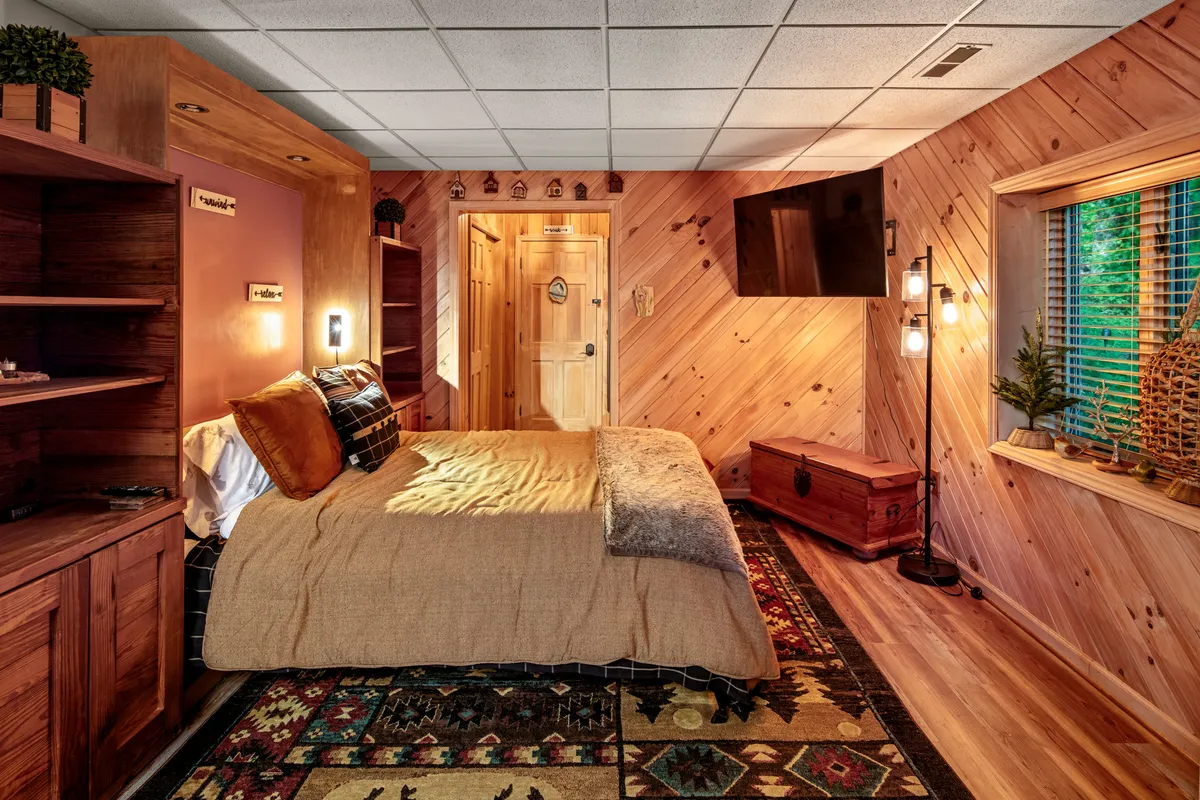 A serene bedroom with a comfortable bed, wooden shelving, TV, decorative trunk, and a window with potted plants. Natural light streams in, creating a cozy space for reading, restful nights, or enjoying morning views. The setup encourages both relaxation and quiet activities like watching TV or reading.