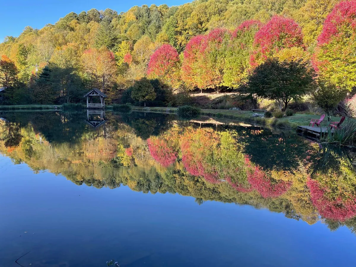 A tranquil pond surrounded by dazzling autumn foliage, a small gazebo, and vibrant red chairs, capturing the beauty of fall and opportunities for fishing, nature walks, and peaceful gatherings.