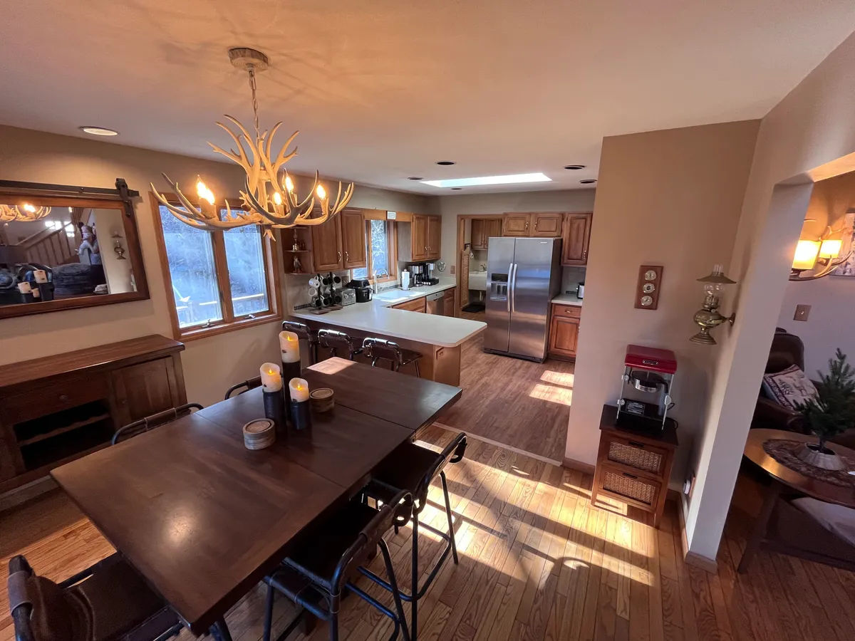 A warm and welcoming dining area with a large table set with candles, a rustic chandelier above, and kitchen appliances and wooden cabinetry in the background. The space feels ready for both festive banquets and casual breakfasts, inviting family and friends to gather around.