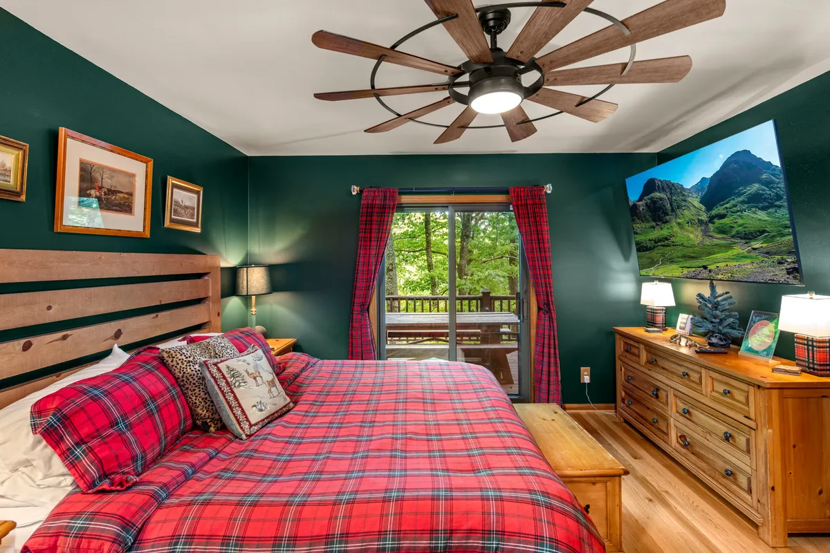 A warmly lit bedroom with plaid bedding, wooden furniture, and a large ceiling fan. The room features framed pictures on the wall and expansive windows that reveal lush natural scenery, filling the space with gentle daylight. The inviting bedding and comfortable décor create a restful atmosphere ideal for relaxation after an active day.