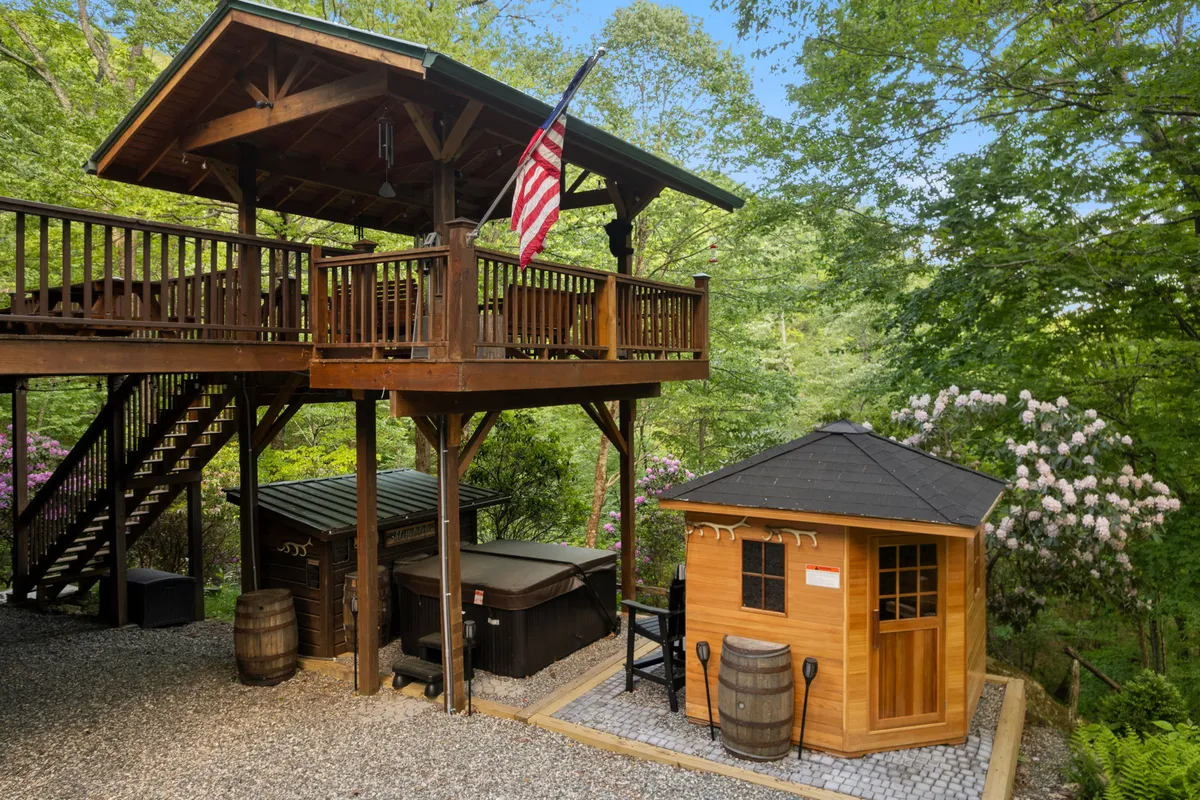 This image shows a spacious wooden deck featuring a private hot tub and a sauna, framed by lush greenery. The American flag and surrounding trees add a sense of place and tranquility. The deck offers space for socializing and relaxing, with open views to the surrounding natural landscape.