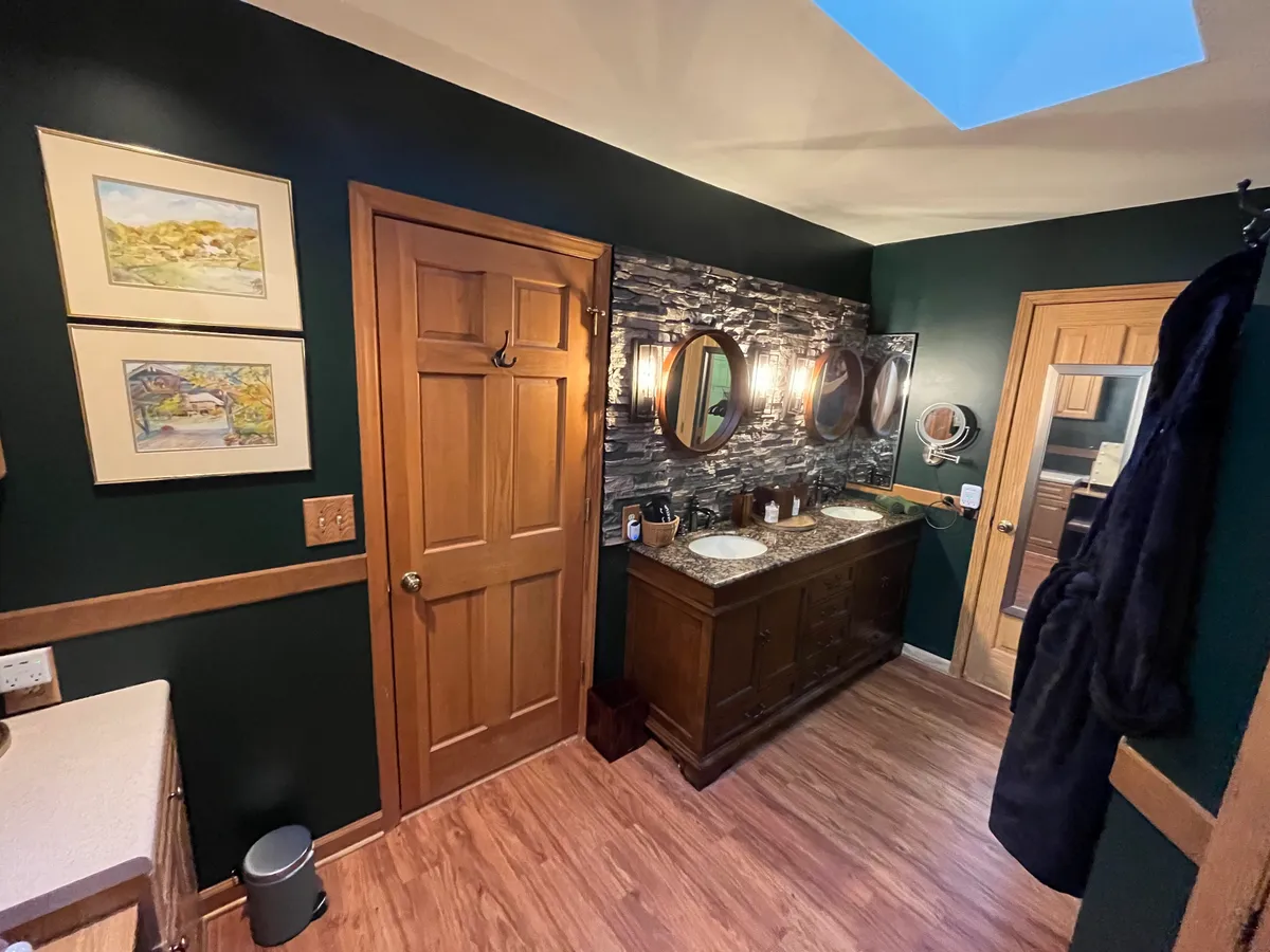 A sophisticated bathroom with a double sink vanity, framed artwork, and mirrors. The wooden door and textured wall add a touch of rustic charm, while the calm lighting invites a sense of relaxation and privacy. The space is meticulously maintained and perfect for personal rituals or unwinding in peace.