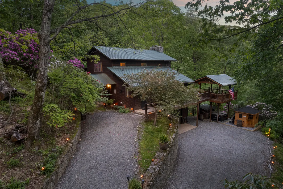 The main house sits nestled among rich greenery, with a welcoming deck featuring a flag. The deck offers expansive views and is set up for gatherings, morning coffee, and relaxation in nature. The house blends seamlessly into its environment, emphasizing seclusion and natural beauty.