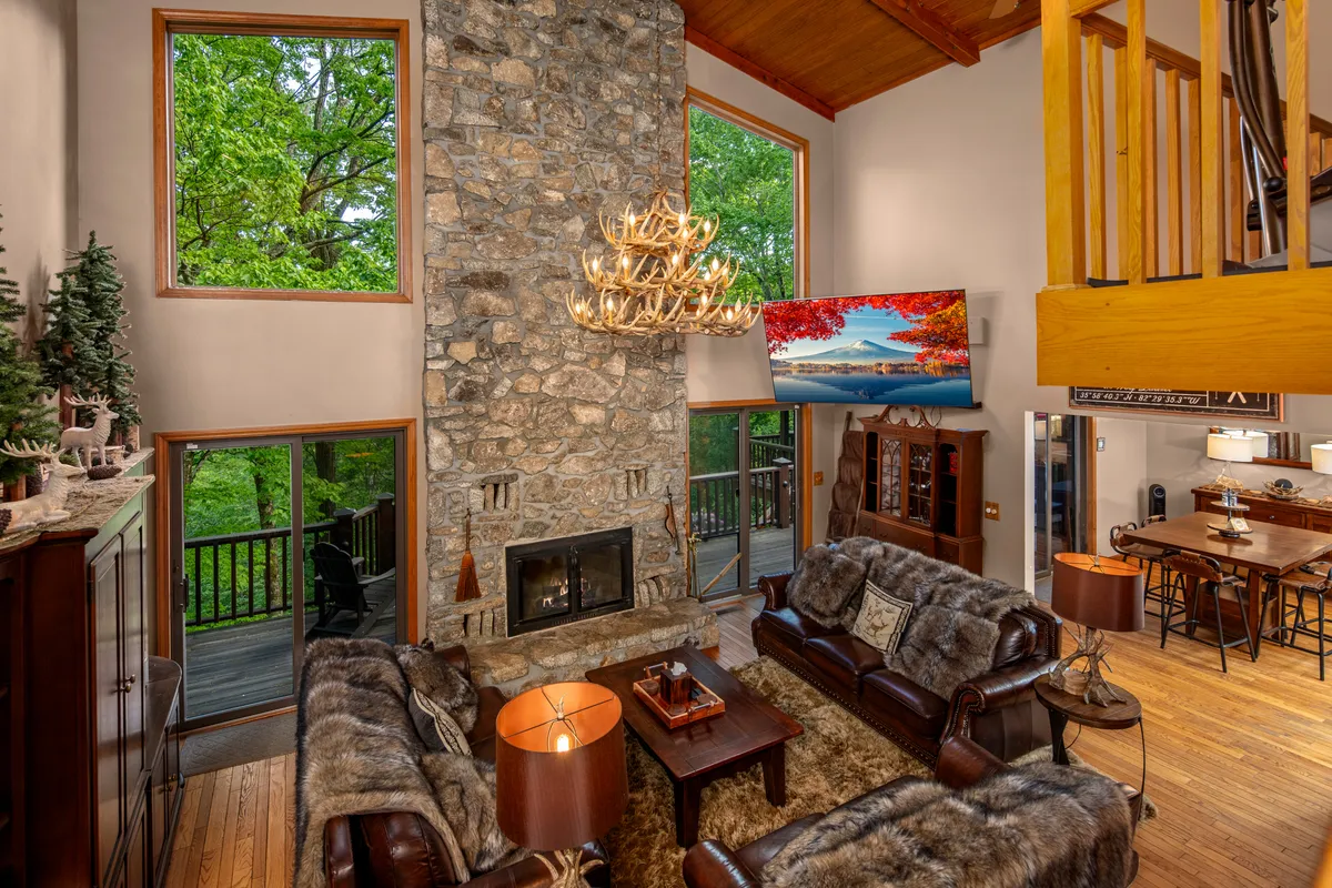 A spacious open-concept living area features a grand stone fireplace, elegant chandelier, and plush leather sofas arranged for conversation and relaxation. Floor-to-ceiling windows invite sunlight and reveal panoramic mountain views, seamlessly connecting the space to the outdoors. The dining area is visible in the background, ensuring flow between gathering spaces.
