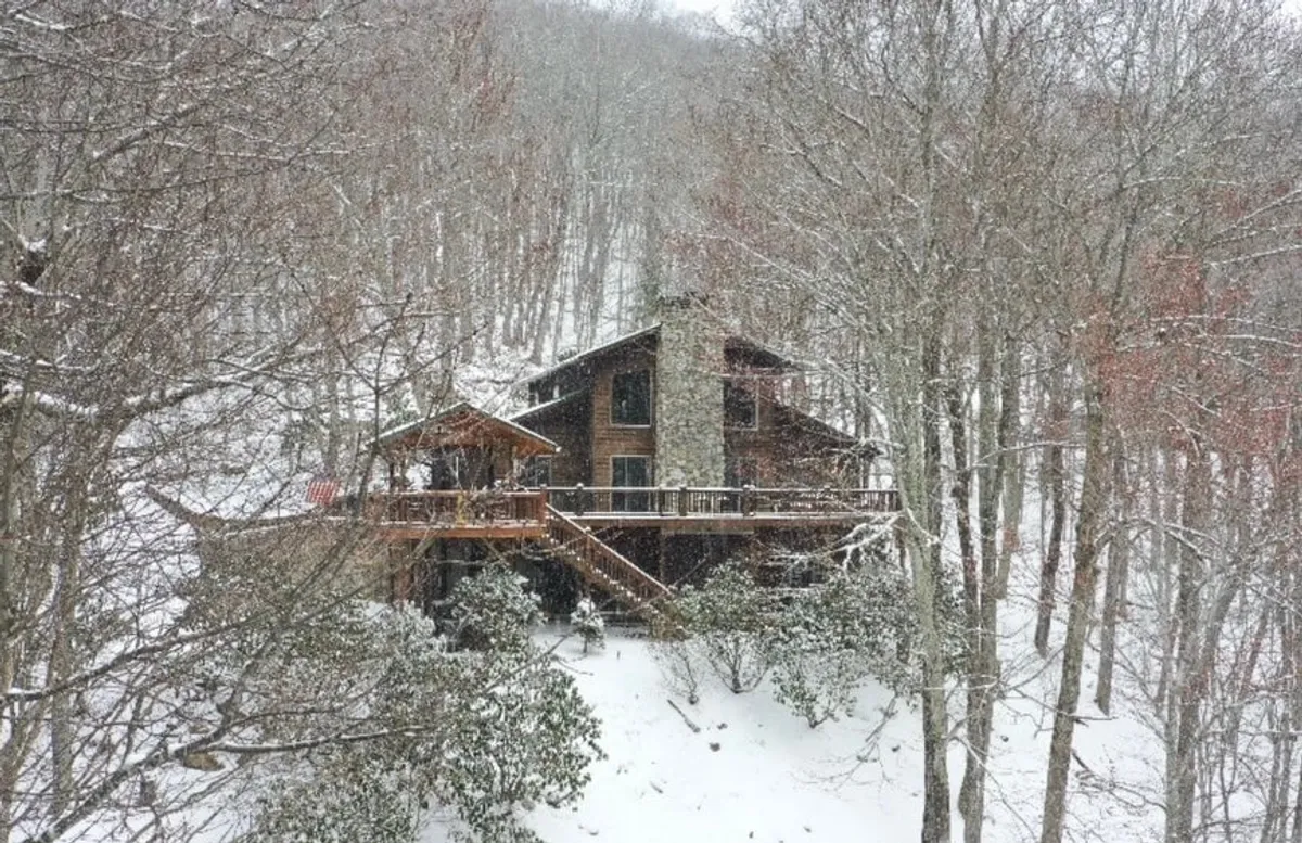 The image showcases the exterior of a spacious mountain lodge, featuring a wooden deck overlooking a snowy landscape. A striking stone chimney hints at a warm fireplace inside, while the surrounding frost-dusted pines create a tranquil winter backdrop. The deck offers a perfect vantage point for watching gentle snowfall or spotting wildlife, and the yard is ideal for snowshoeing or building snowmen.