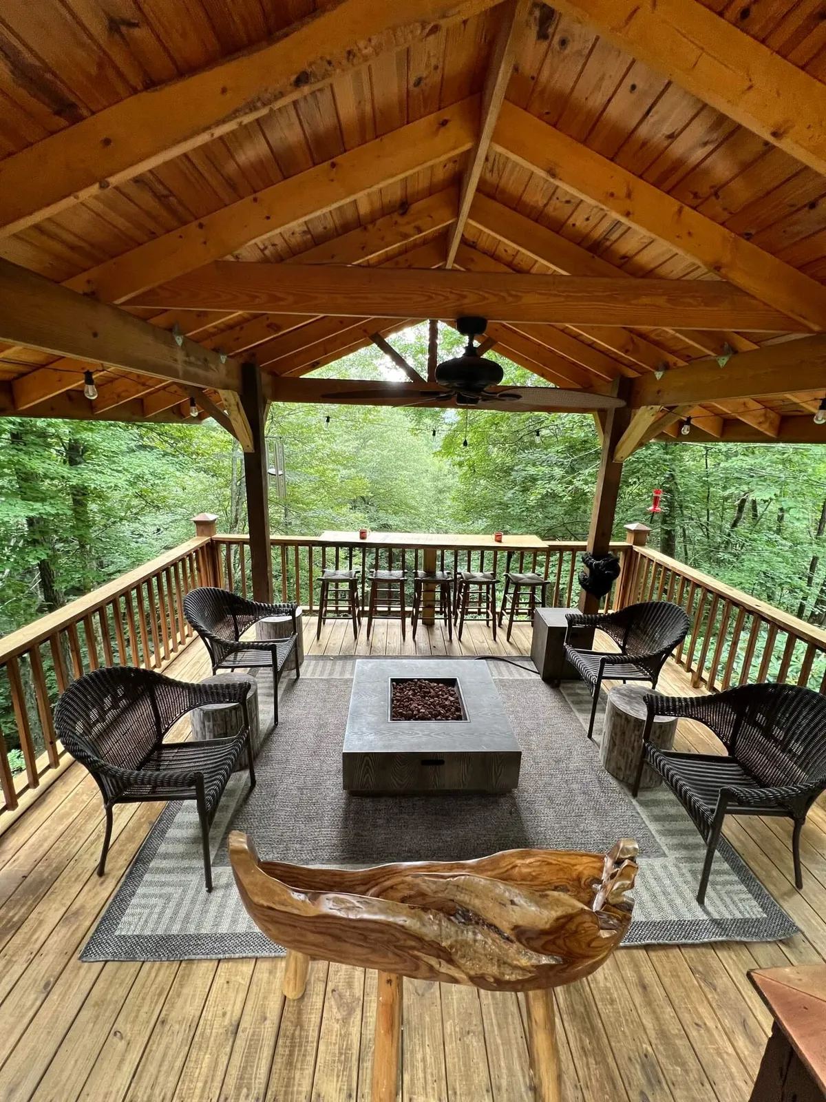 A cozy exterior deck with a fire pit, wooden ceiling, wicker chairs, and a dining area in the background. Surrounded by scenic greenery, this space is set up for social gatherings, coffee at sunrise, or evening stargazing by the fire.