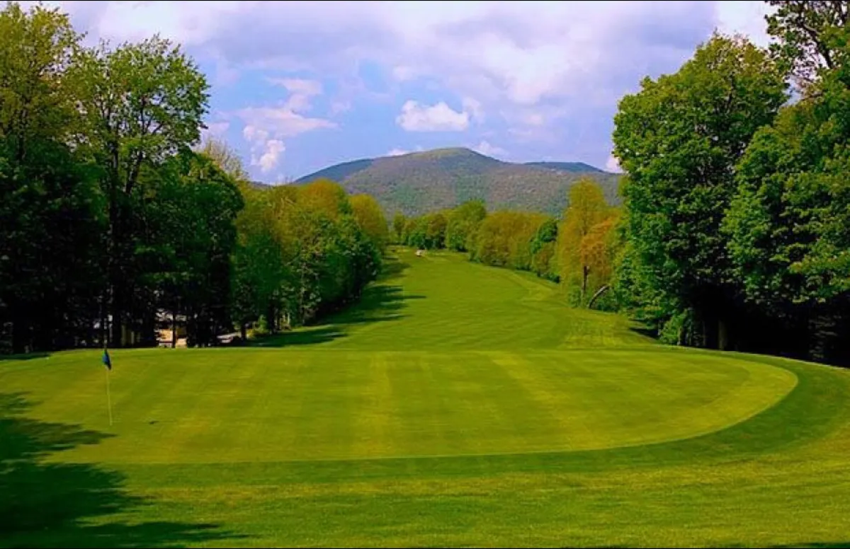 This image showcases a lush golf course fairway set against a stunning mountain backdrop under a clear blue sky. The scene captures the essence of country club leisure, with expansive green lawns and dramatic natural vistas that invite visitors to relax or partake in outdoor recreation.